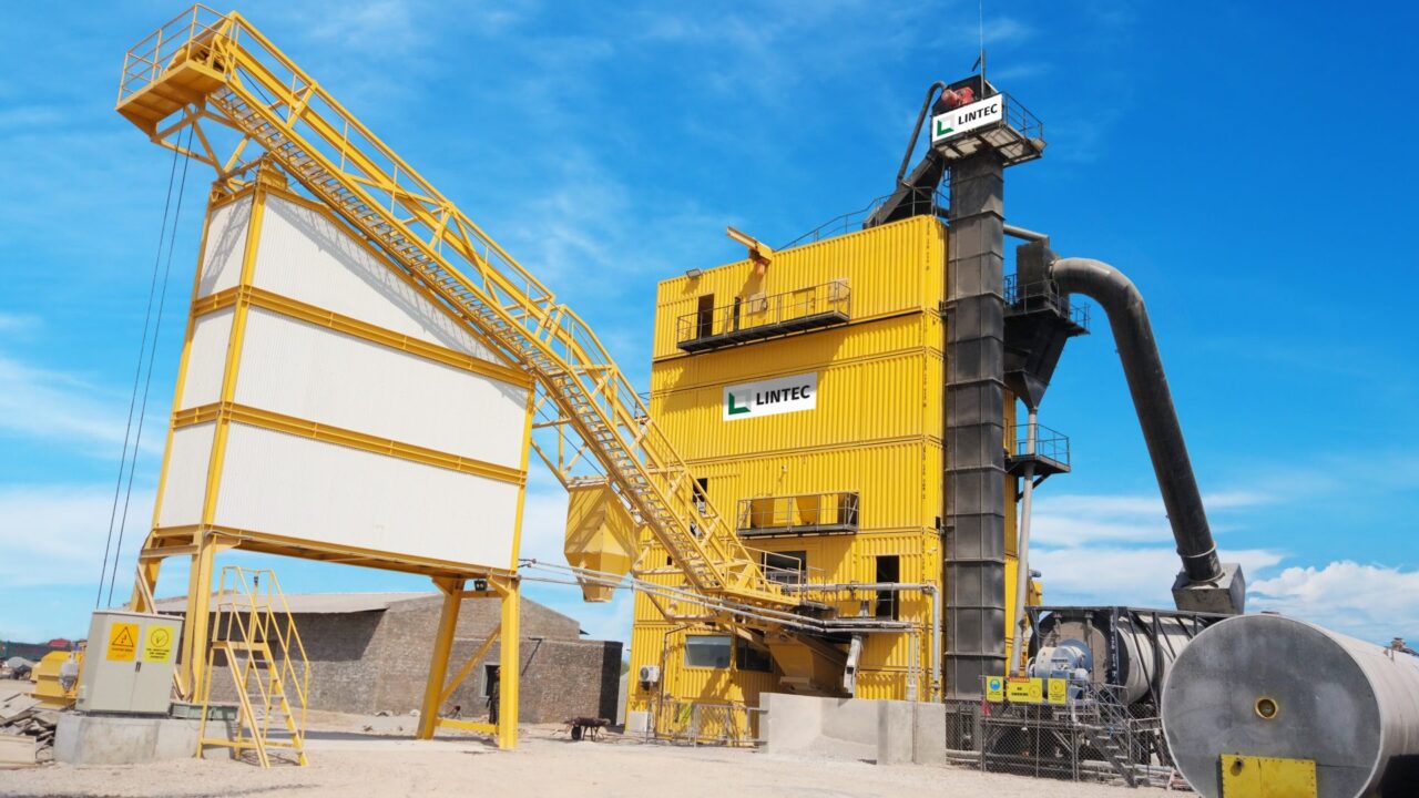 A yellow Lintec containerized asphalt batching plant with a modular structure and conveyor system at a road construction site.