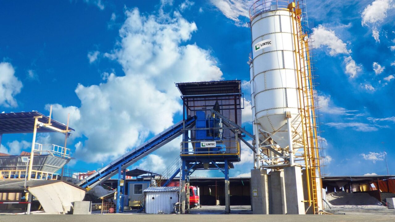 A Lintec concrete batching plant featuring a tall white cement silo and an inclined conveyor belt feeding the central mixing unit.